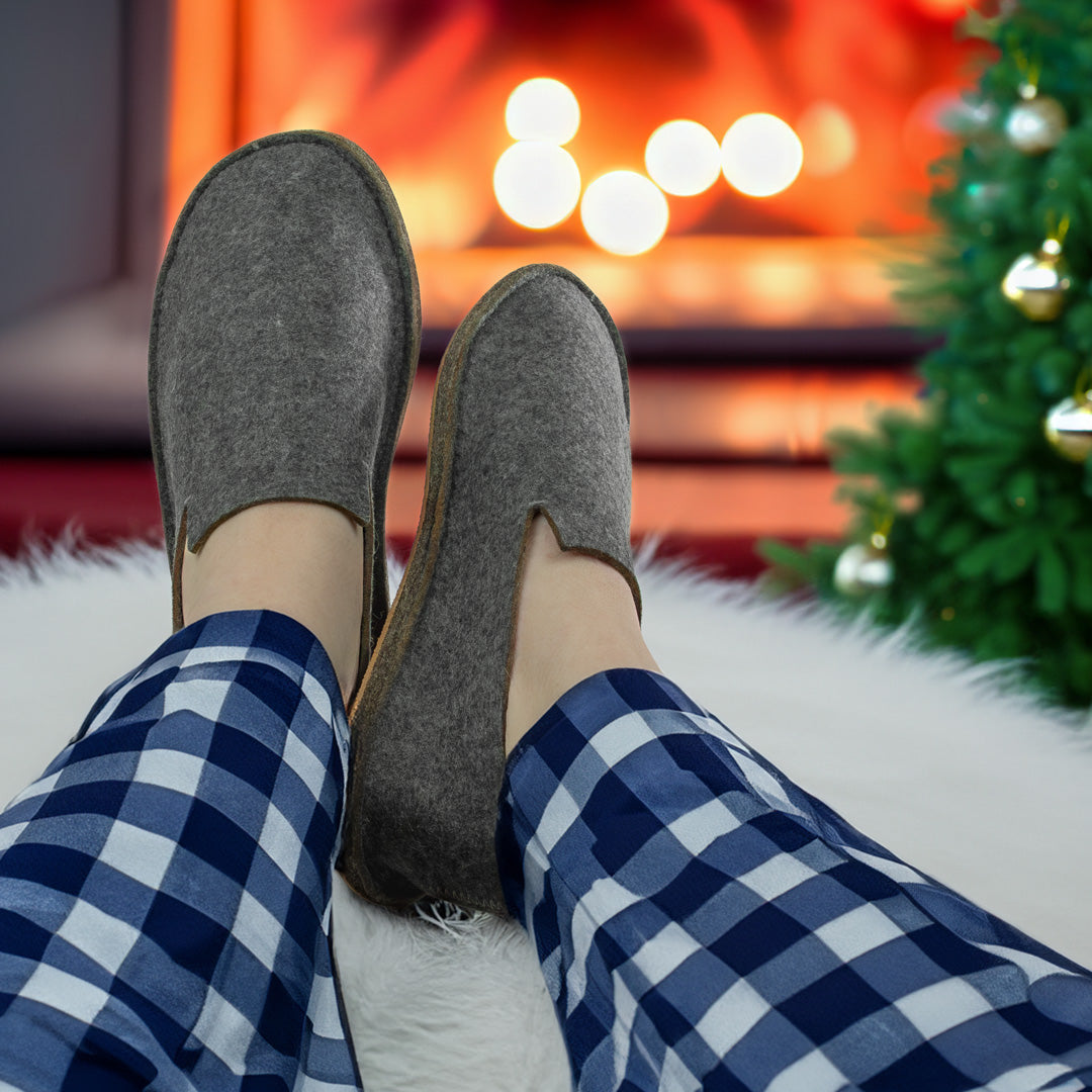 Cozy holiday scene with a person in blue plaid pajamas wearing The Felt Store's 100% Wool Felt Slippers — A warm ambiance is set by a wood-burning fireplace and a decorated Christmas tree — The perfect gift for him and her